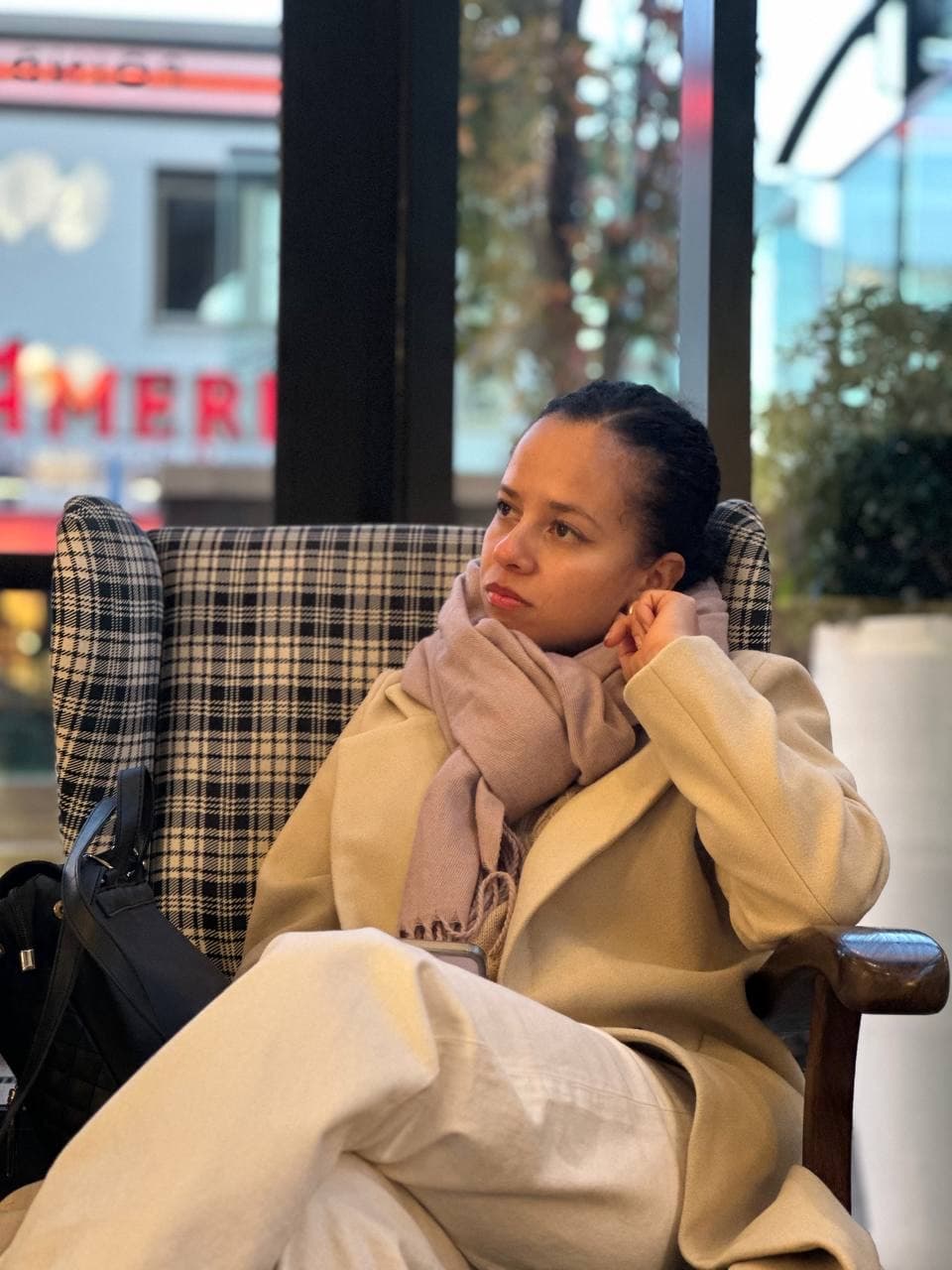 Cami sitting thoughtfully in a checkered armchair, wearing a cream coat